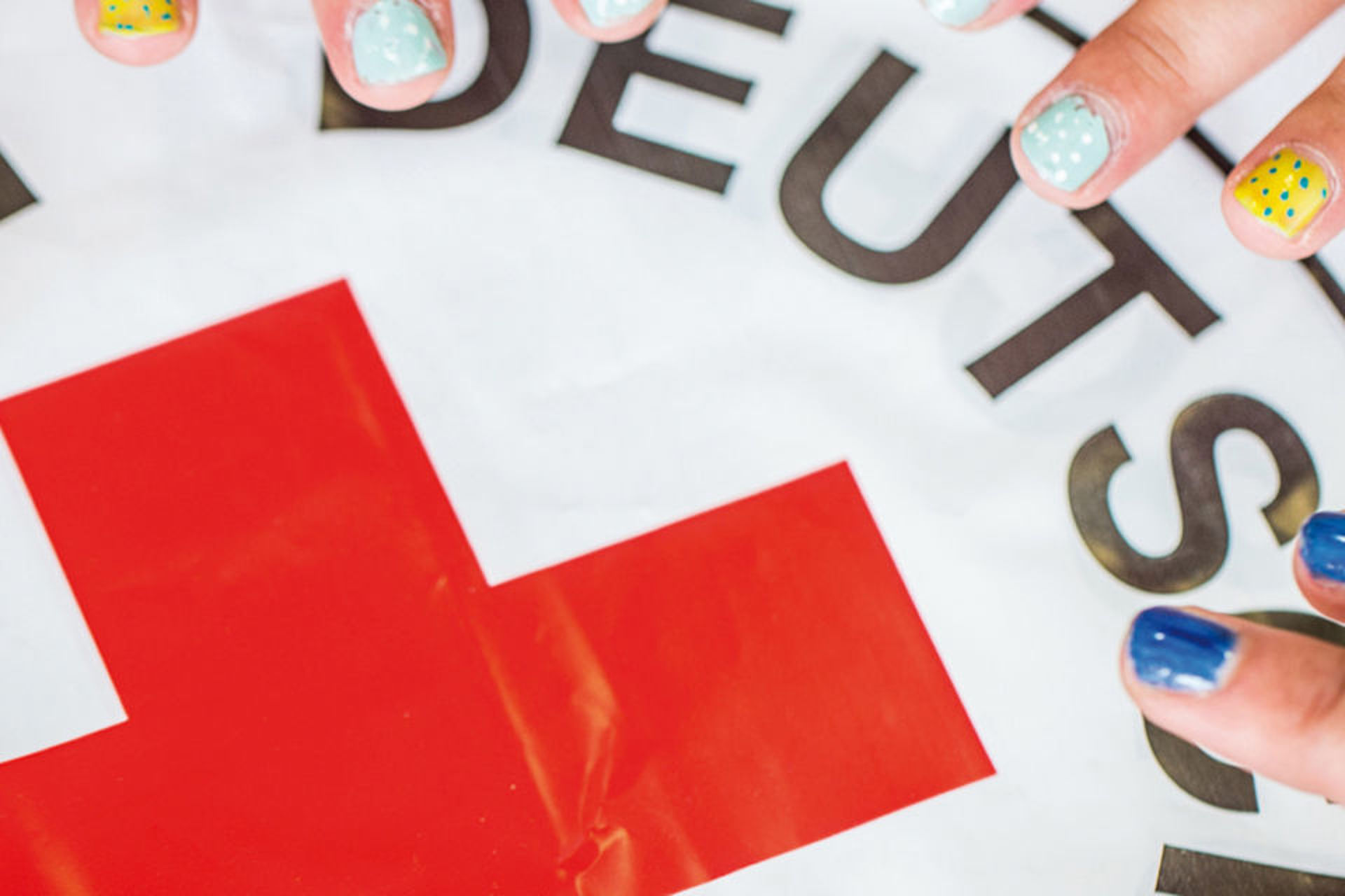 DRK Logo, auf dem mehrere Hände mit bunt lackierten Nägel liegen.
