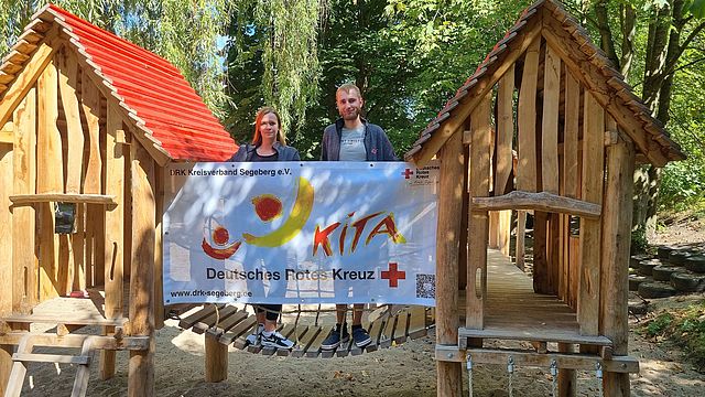 Zwei DRK-Mitarbeitende stehen auf einem Kinderspielplatz vor einem großen Banner mit dem DRK Kita Logo.