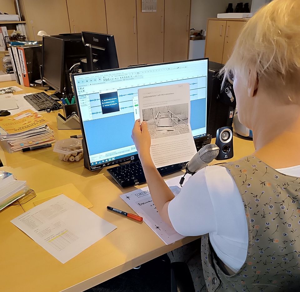 Foto einer Person in Rückenansicht, die vor einem Mikrofon eine Zeitung vorliest.
