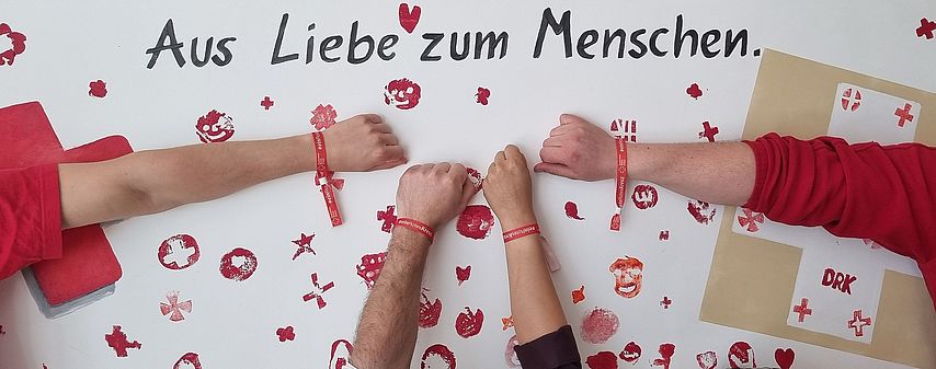 Vier Menschen halten ihre Arme an eine Wand mit der Aufschrift "Aus Liebe zum Menschen". Alle tragen DRK Armbänder und rote DRK Kleidung.