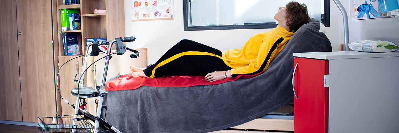 Übungspuppe liegt auf einem Krankenbett. Nebenan steht ein Rollator.