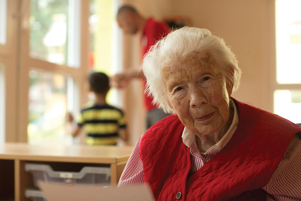demenz Lächelnde ältere Dame liest einen Brief. Im Hintergund steht ein DRK Mitarbeiter mit einem Kind.