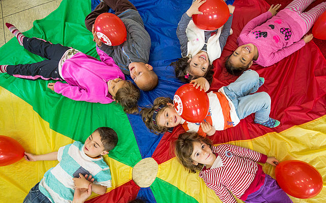 Kinder liegen auf einem bunten Tuch und spielen mit DRK Luftballons.
