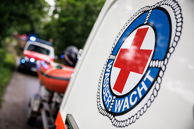 Logo der Wasserwacht auf einem Boot mit einem Rettungswagen im Hintergrund.