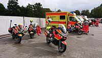 Mehrere Motorräder der DRK Motorradstaffel stehen neben einem DRK Rettungswagen. Im Hintergrund befindet sich eine Gruppe von Menschen in DRK Bereitschaftskleidung.