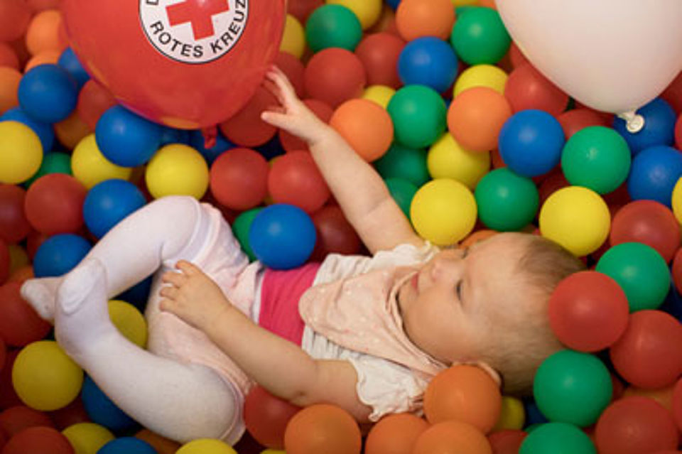 Foto eines Babys, dass in einem bunten Bällebad liegt. Ein roter DRK-Luftballon liegt neben dem Baby.