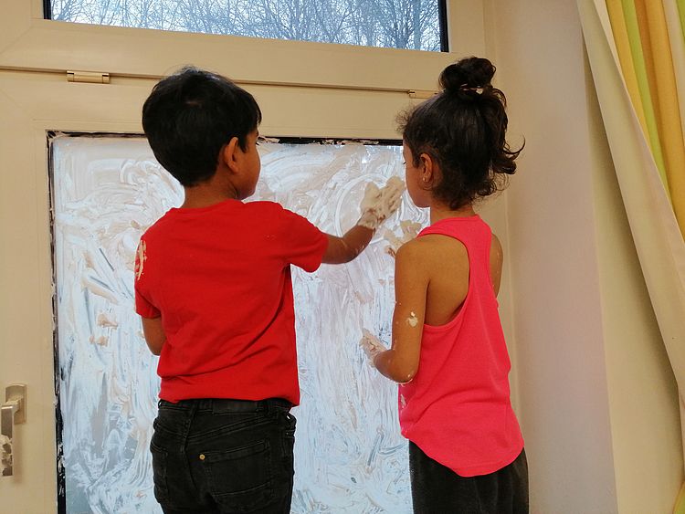 Zwei Kinder spielen mit Schaum an einem Fenster.
