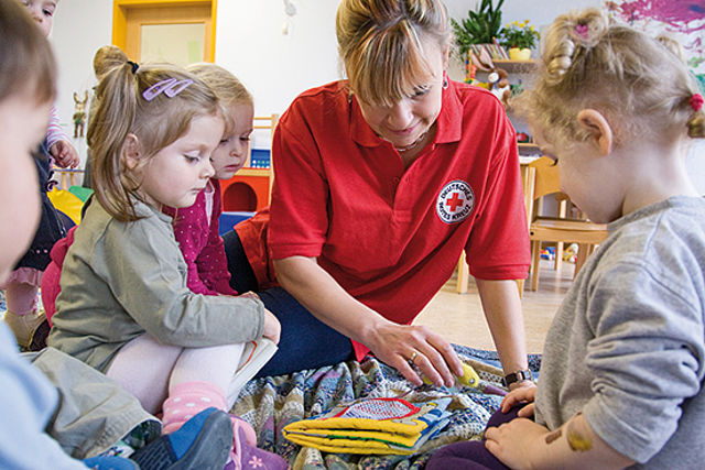 Eine DRK-Mitarbeiterin, in rotem DRK-Shirt, spielt mit mehreren Kita-Kindern auf dem Boden.