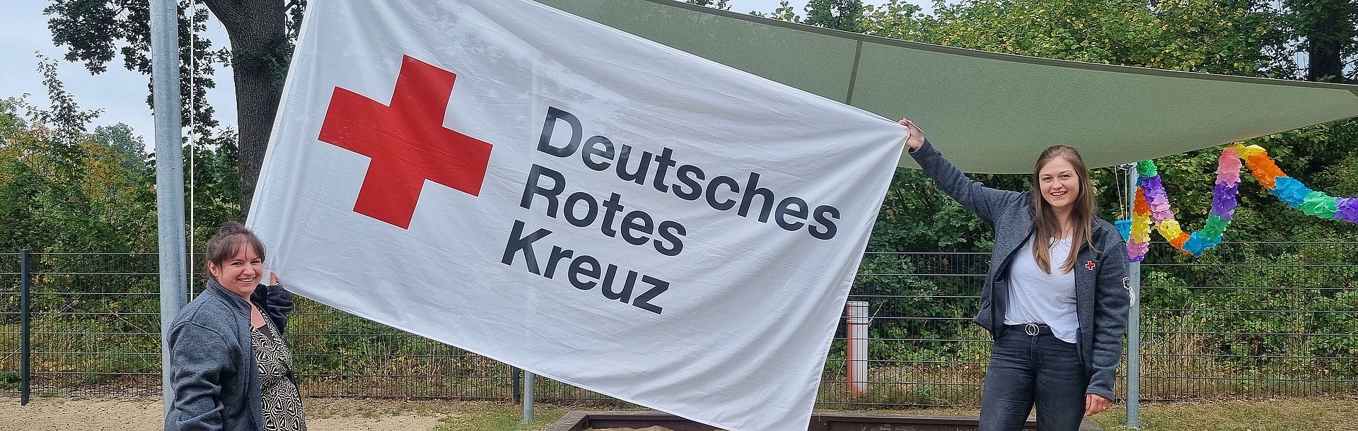 Foto von zwei DRK-Mitarbeiterinnen, die eine große Deutsches Rotes Kreuz Flagge zwischen sich halten.