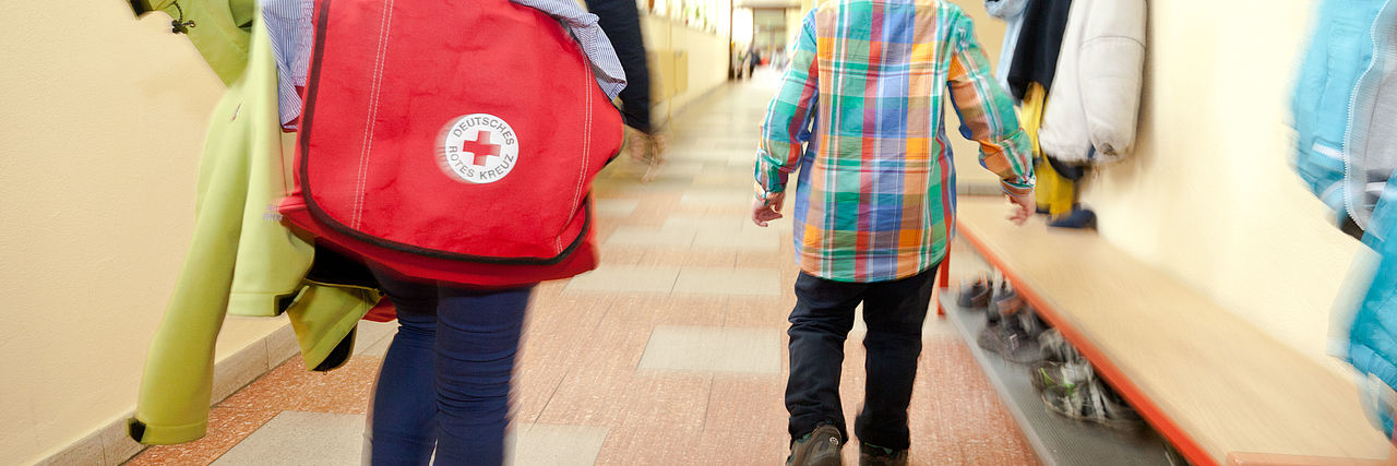 Ein Erwachsener mit DRK Tasche geht neben einem Kind in eine Schule.