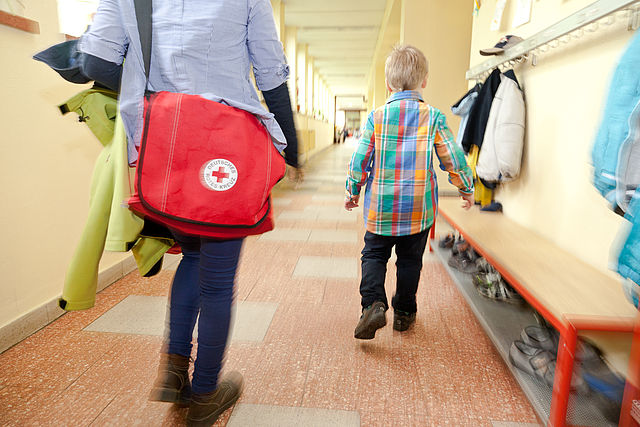 Ein Erwachsener mit DRK Tasche geht neben einem Kind in eine Schule.