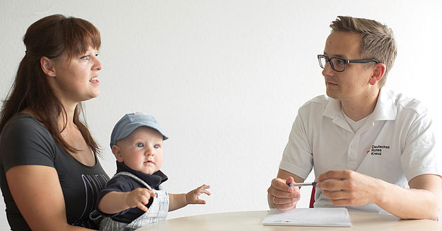 Foto eines DRK Mitarbeiters, der mit einer Mutter und ihrem Kleinkind spricht.