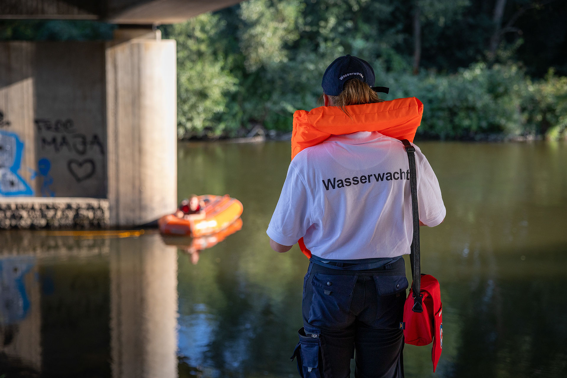 Mitarbeiterin der Wasserwacht mit einer Rettungsweste steht vor einem Fluss, in welchem ein Rettungsbot schwimmt.