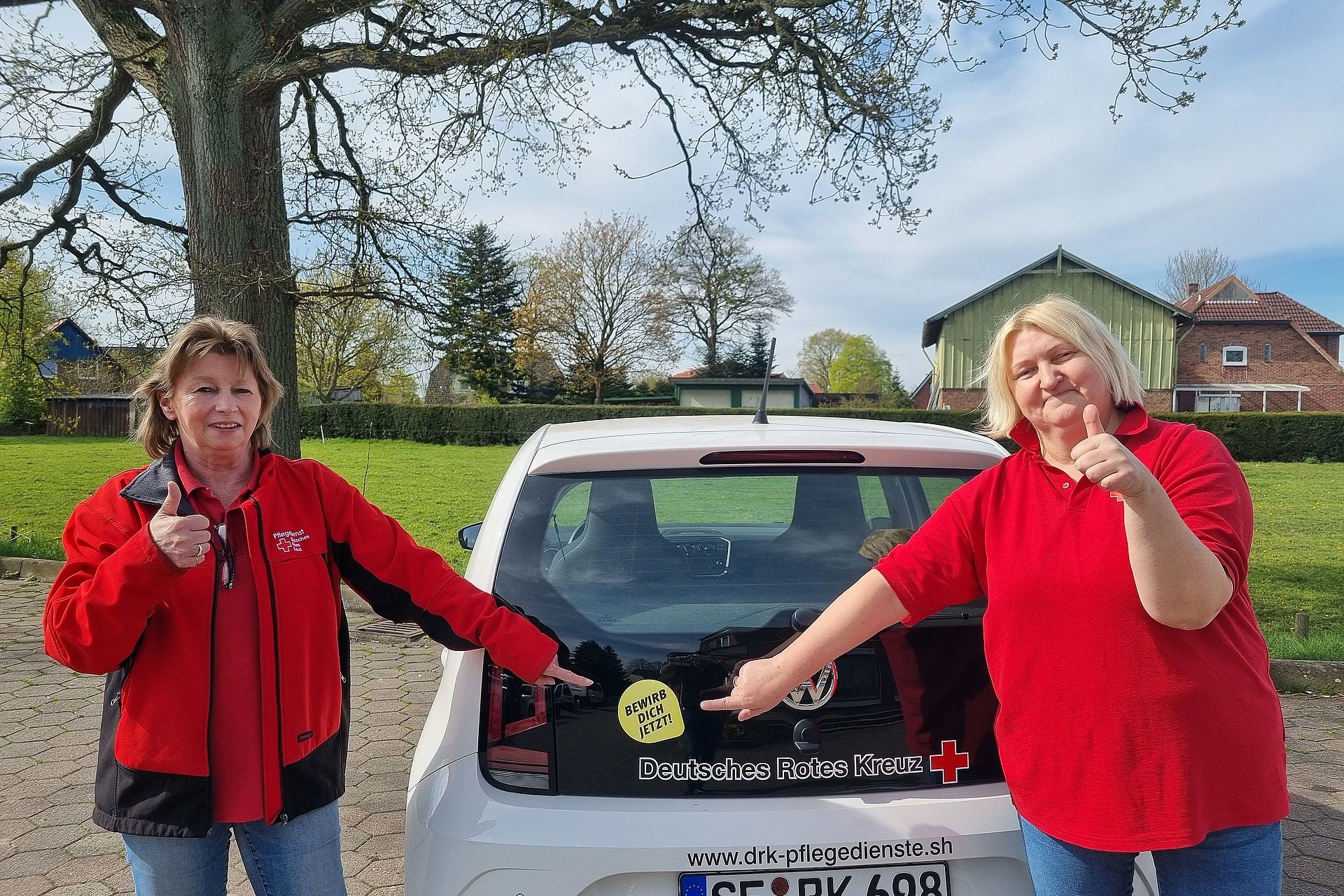 Foto von zwei DRK Mitarbeiterinnen, die in roter DRK Kleidung auf einen "Bewirb dich jetzt"-Aufkleber zeigen.