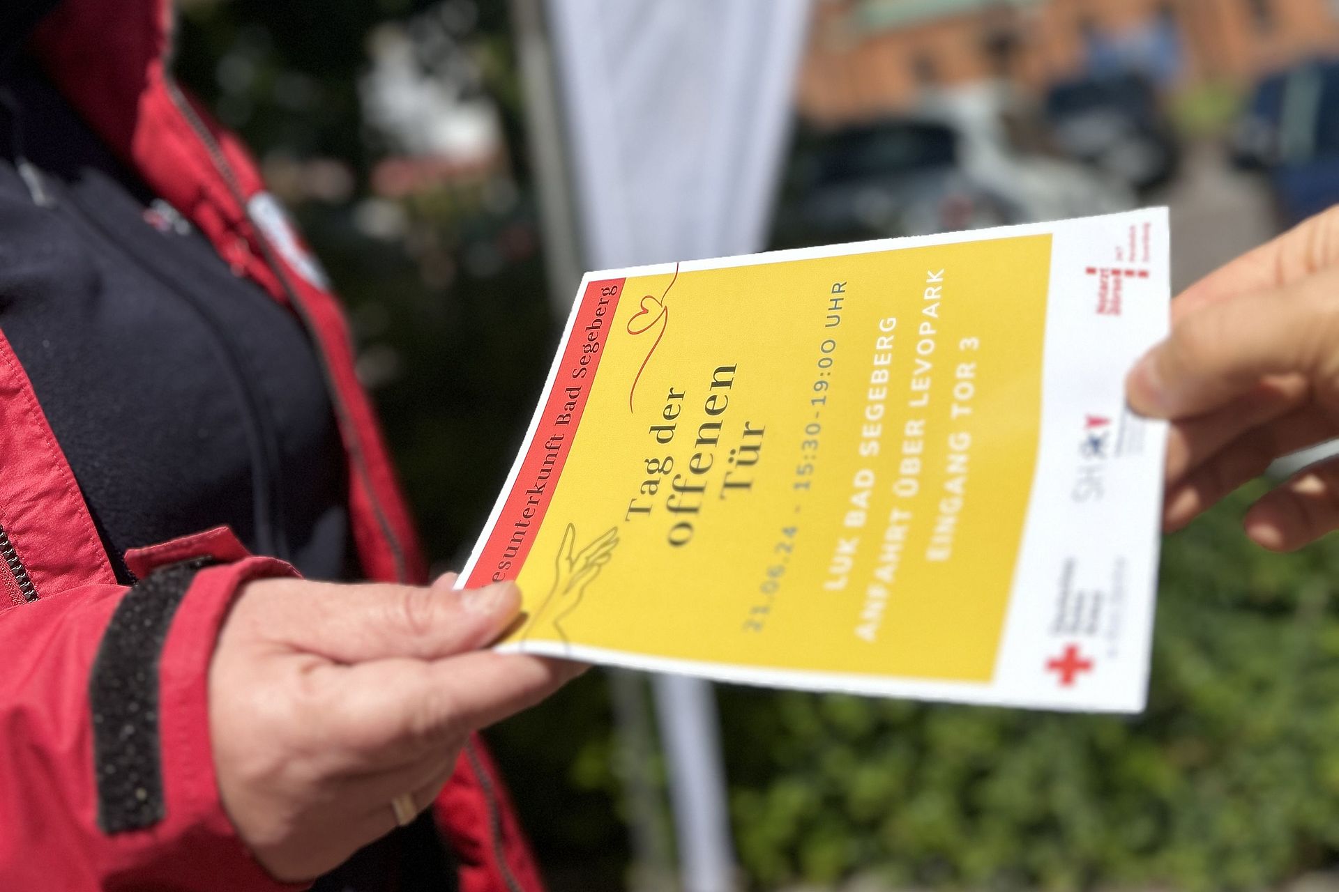 DRK Mitarbeiterin gibt einer anderen Person einen Flyer "Tag der offenen Tür:  DRK Landeunterkünfte Bad Segeberg"