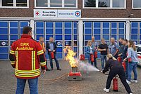 Mehrere Personen stehen um ein Feuer und eine Person löscht dieses mit einem Feuerlöscher.